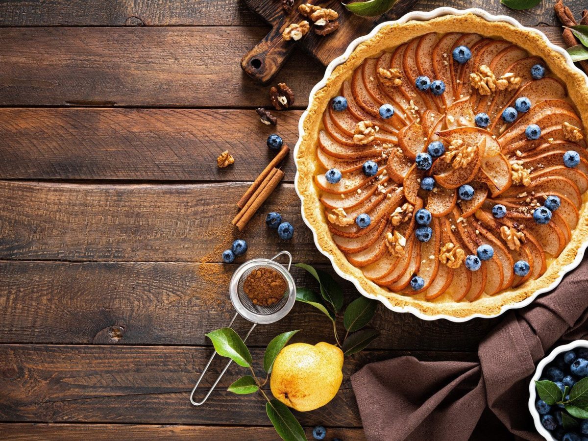 Auf einem dunklem Holztisch steht eine vegane Birnen-Walnuss-Tarte. Sie ist mit gemahlenen Mandeln und Blaubeeren dekoriert.