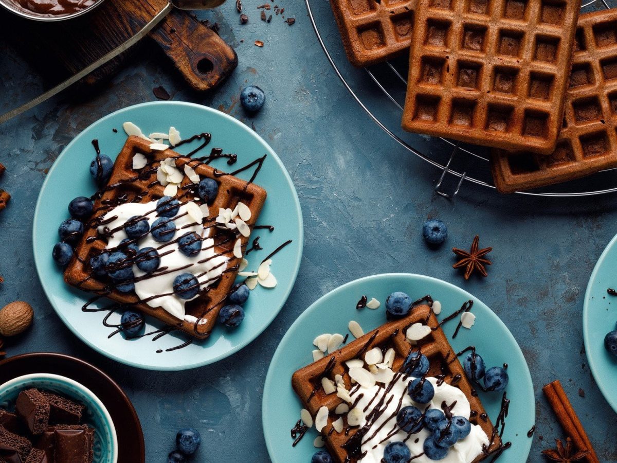 Auf drei blauen Tellern sind schokoladige Brownie Waffeln verteilt. Sie sind dekoriert mit Blaubeeren, Naturjoghurt und Schokoladensauce.