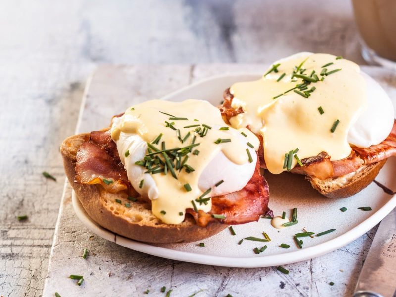 Eggs Benedict mit Speck auf Brot und Hollandaise, angerichtet auf einem weißen Teller.