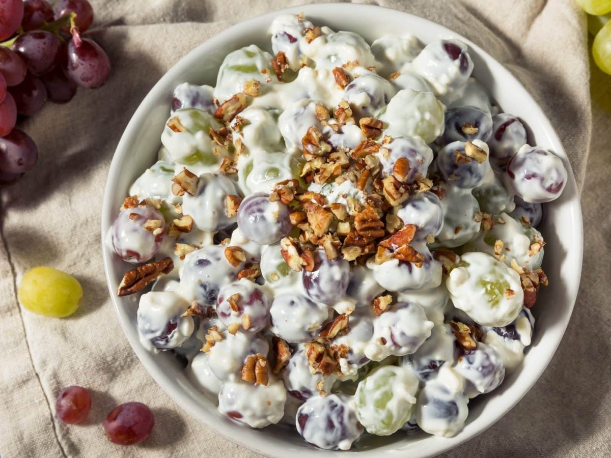 Der cremige Traubensalat mit Nüssen angerichtet in einer weißen Schale. Rundherum liegen rote und weiße Weintrauben.