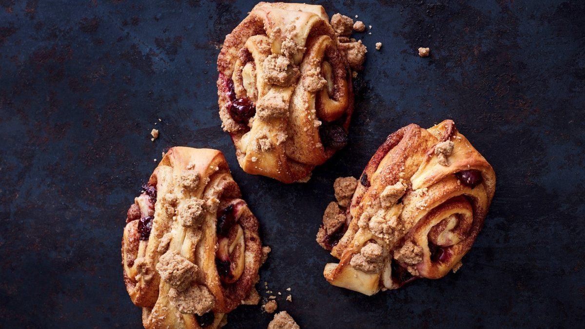 Auf einer dunkelblauen Arbeitsfläche liegen drei Kirsch-Karamell-Franzbrötchen mit Streuseln.