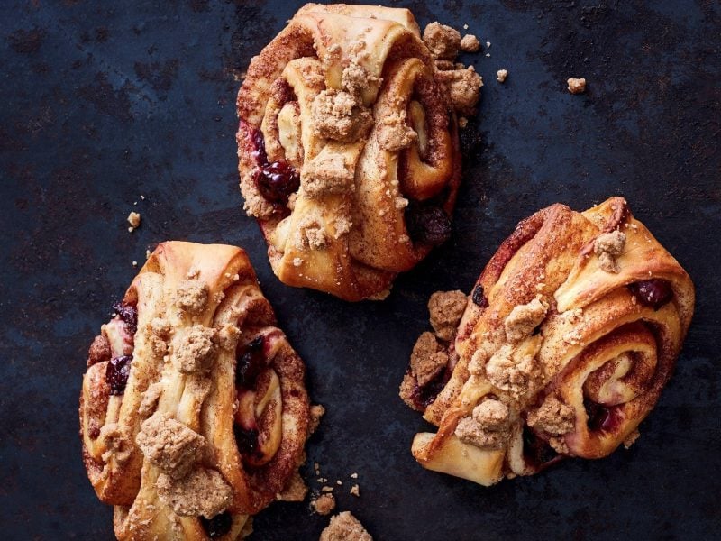 Auf einer dunkelblauen Arbeitsfläche liegen drei Kirsch-Karamell-Franzbrötchen mit Streuseln.