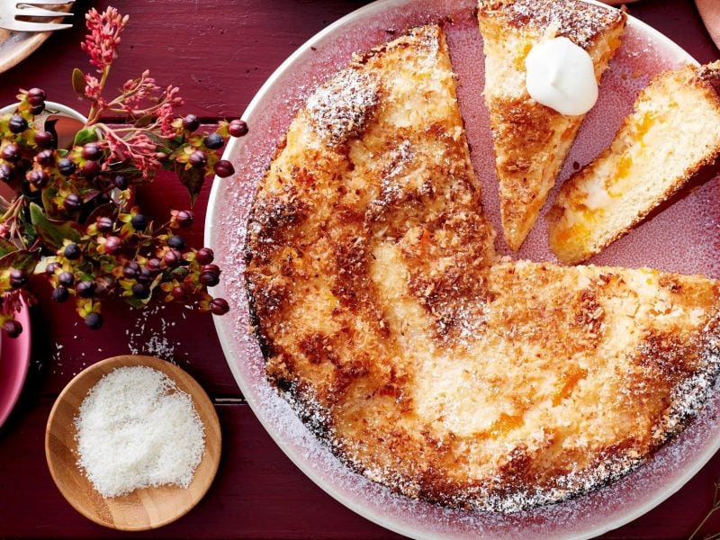 Auf einem Teller liegt ein fluffiger Mandarinen-Kokos-Kuchen. Er ist mit Puderzucker bestreut und mit einem Klecks Schlagsahne verfeinert.