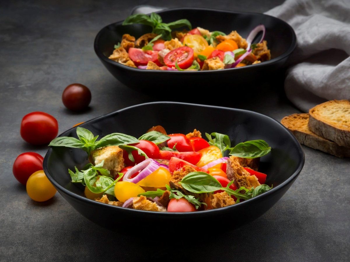 Zwei Teller befüllt mit italienischem Brotsalat Panzanella auf dunklem Untergrund.