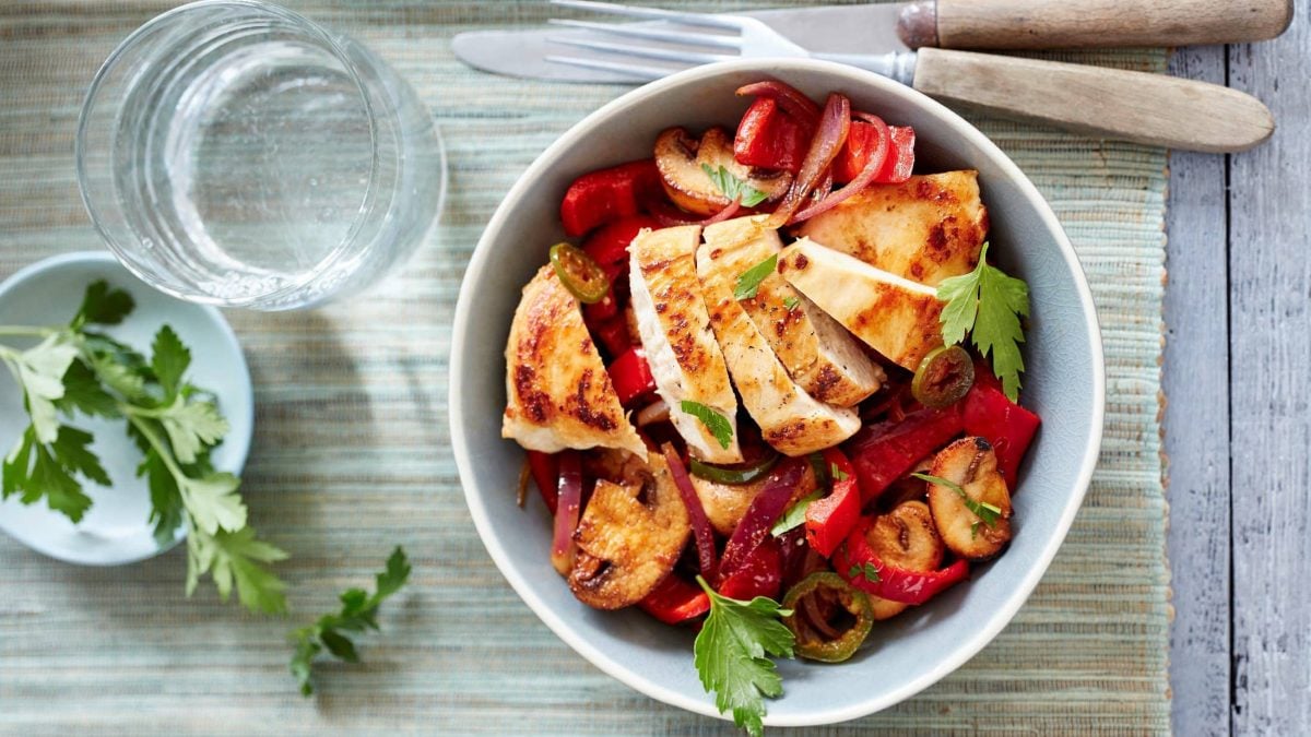 In einer weißen Porzellan-Schüssel ist eine Paprika-Pilz-Pfanne mit Huhn zubereitet. Daneben liegen Messer und Gabel