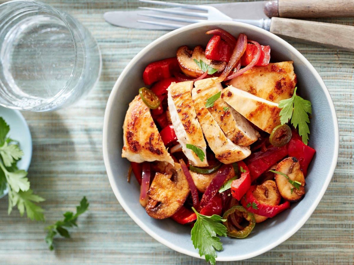 In einer weißen Porzellan-Schüssel ist eine Paprika-Pilz-Pfanne mit Huhn zubereitet. Daneben liegen Messer und Gabel