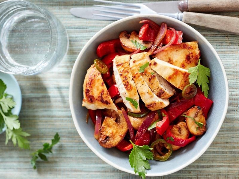 In einer weißen Porzellan-Schüssel ist eine Paprika-Pilz-Pfanne mit Huhn zubereitet. Daneben liegen Messer und Gabel