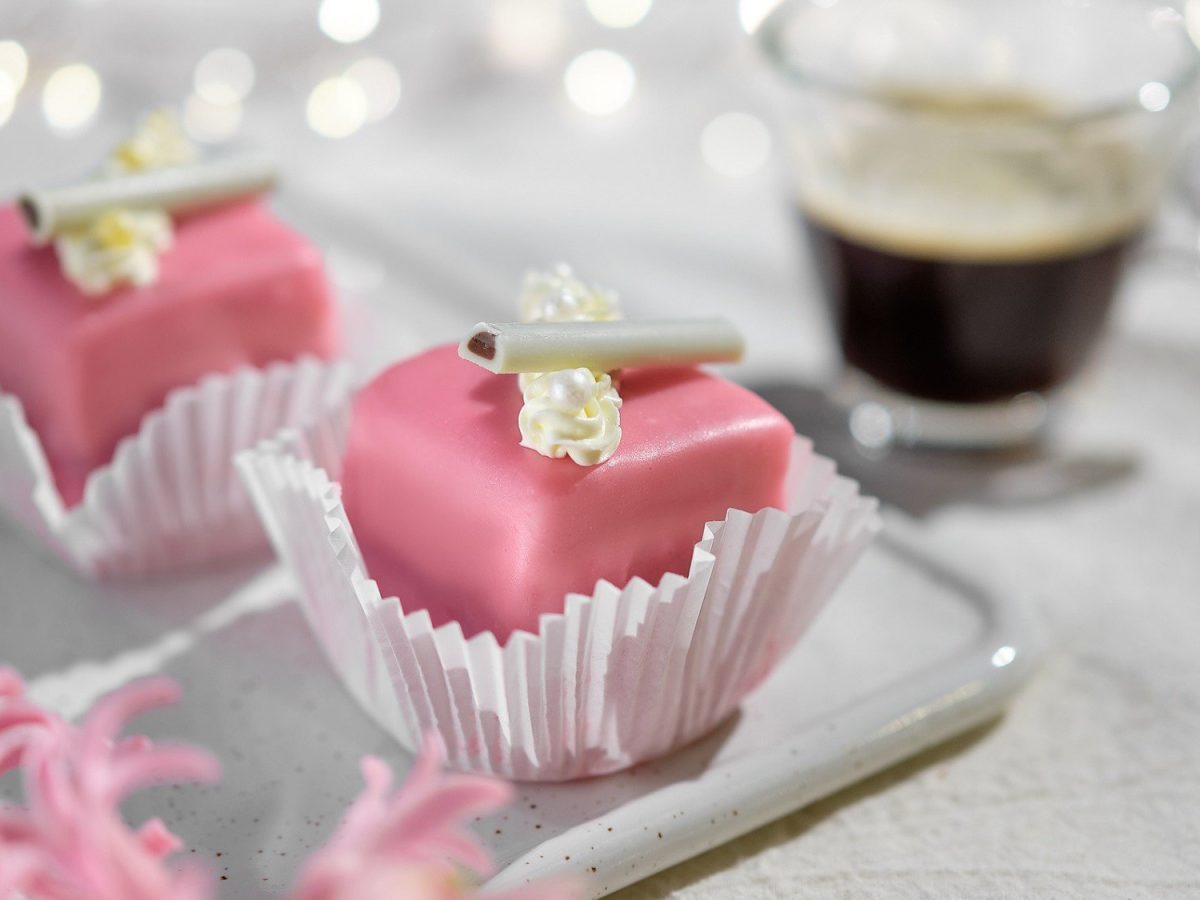 In einer kleinen Papierform sind Petit Fours angerichtet. Sie sind mit rosa Marzipan überzogen und mit Zuckerperlen dekoriert.