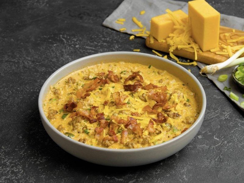 Die Low-Carb-Cheeseburger-Pfanne angerichtet in einem weißen Teller. Im Hintergrund geriebener Käse und Frühlingszwiebeln.