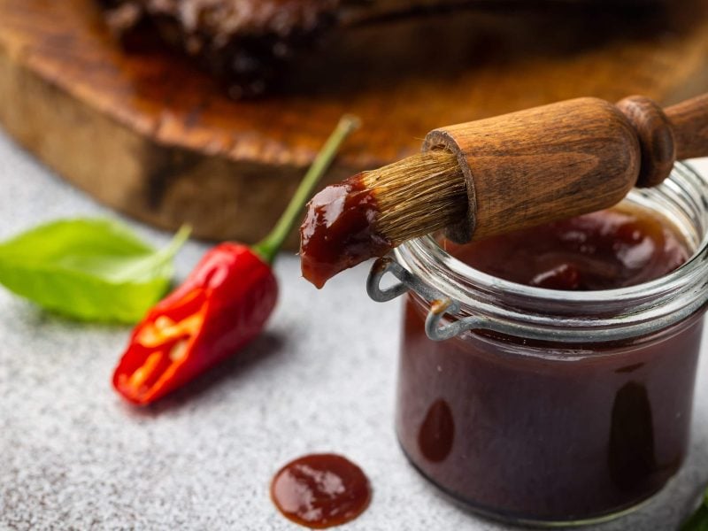 Basting Marinade und Sauce im Glas mit Glasur-Pinsel. Chili und Fleisch auf einem Holzbrett im Hintergrund. Frontalansicht.