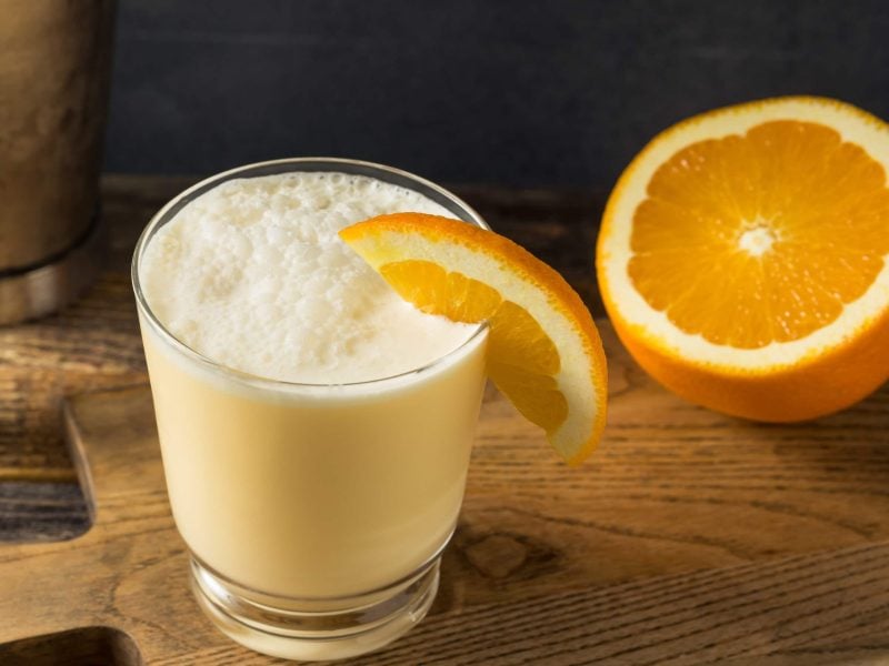 Orange Whip aus "Blues Brothers" in Longdrinkglas mit Orangenschale auf Holztisch. Daneben rechte eine halbe Ornae mit der Schnittseite zum Betrachter. Link Cocktailzubehör. Aufnahme aus der Frontalen.