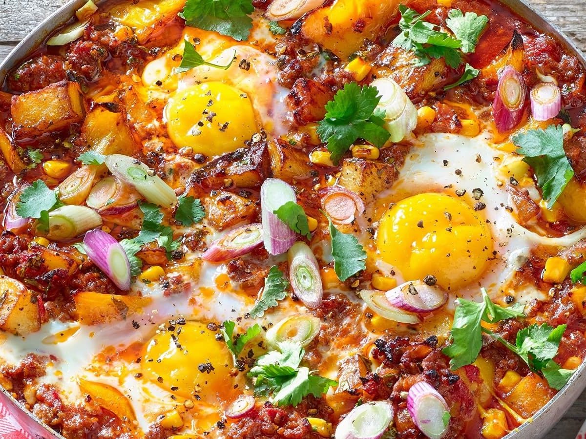 Eine große Pfanne mit dem Bolognese-Shakshuka, garniert mit Korianderblättern und Frühlingszwiebeln, bestreut mit frisch gemahlenem Pfeffer neben einem rot-weiß karierten Geschirrtuch und einem Teller mit einem Stück Brot, vor einem Holzhintergrund von oben.