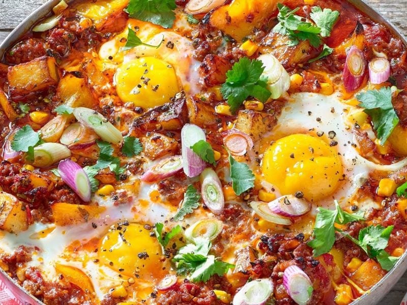 Eine große Pfanne mit dem Bolognese-Shakshuka, garniert mit Korianderblättern und Frühlingszwiebeln, bestreut mit frisch gemahlenem Pfeffer neben einem rot-weiß karierten Geschirrtuch und einem Teller mit einem Stück Brot, vor einem Holzhintergrund von oben.