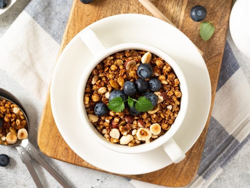 Eine Schale Buchweizenmüsli mit Haselnüssen und Blaubeeren in der Draufsicht, daneben ein Löffel Müsli und ein Kännchen Milch.