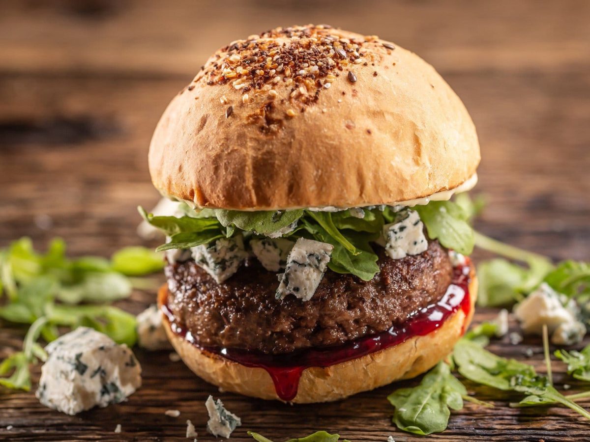 Ein Burger mit Blauschimmelkäse und Preiselbeersauce auf einem Untergrund aus dunklem Holz. Um den Burger herum liegen noch Rucola und Blauschimmelkäse.