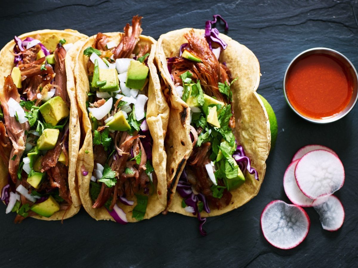 Dreimal Carnitas fuer Tacos Tacos mit Füllung auf schwarzem Tisch. Daneben Radieschen und eine Schüssel mit roter Sauce. Aufnahme aus der Vogelperspektive.