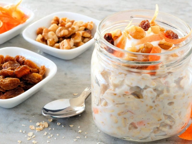 EIn Glas Carrot Cake Overnight Oats, daneben stehen Schälchen mit Zutaten wie Nüssen oder Rosinen, außerdem geraspelte Möhren.