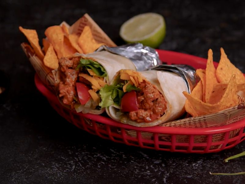 Cheeseburger-Burrito in aufgeschnitten in Alufolie gewickelt mit Nachos in rotem Brotkorb. Schnittseit zum Betrachter. Rechts eine grüne und eine rote Chili. Halbe Limette oben und Dip links im Bild. Frontalaufnahme.