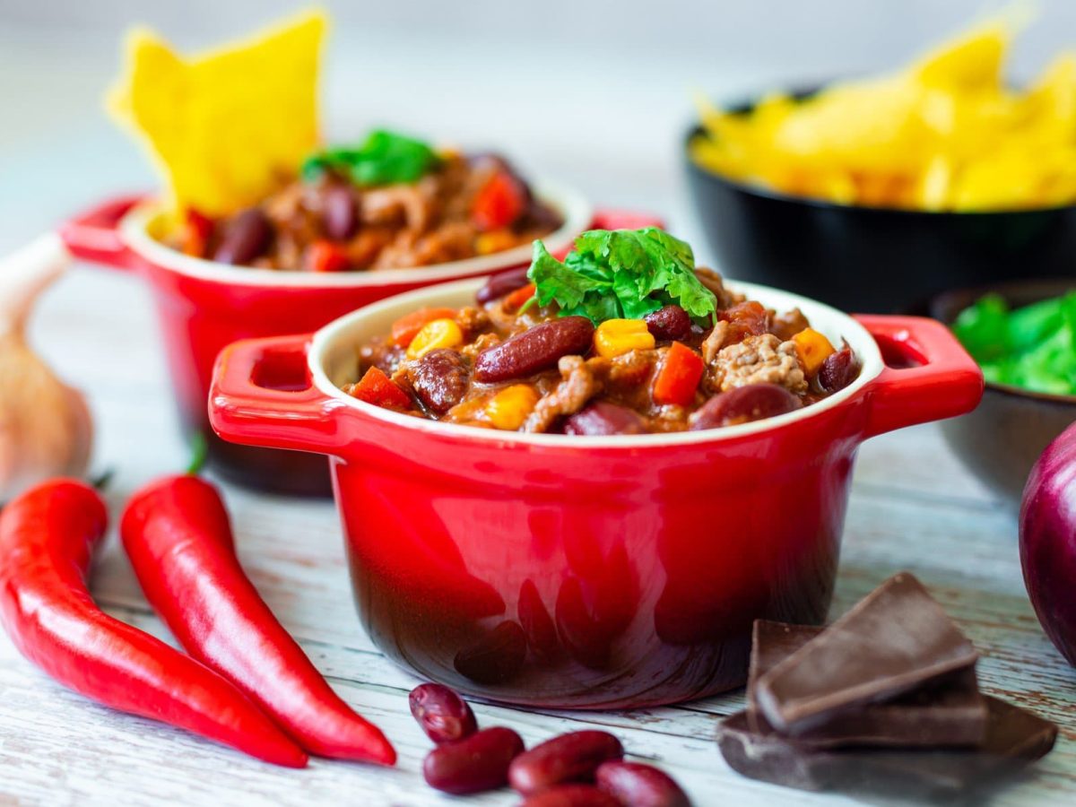 Zwei kleine rote Töpfe Chili sin Carne mit Schokolade, daneben liegen Zutaten wie Chilischoten und Zwiebeln, außerdem ist eine Schale mit Nachos zu sehen.