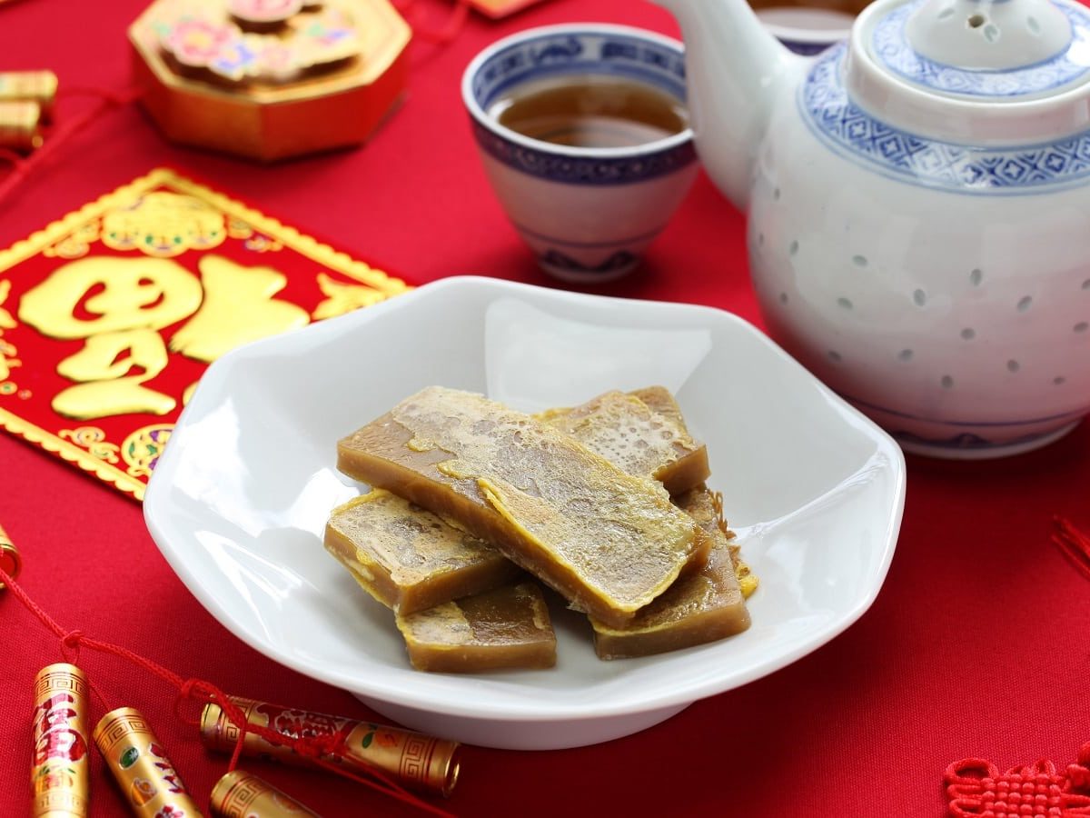 Eine weiße Porzellan-Schüssel mit mehreren Scheiben des chinesischen Reiskuchens Nian Gao neben einer Kanne und einer Tasse mit dunklem Tee, alles auf einem Tisch, der mit einem roten Tuch ausgelegt ist und mit festlichen Dekorationen verziert ist.