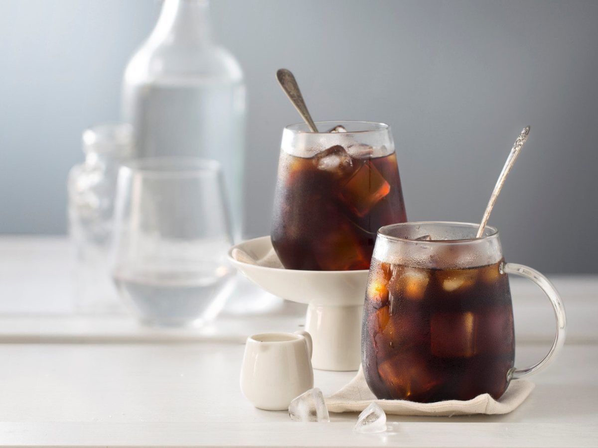 Zwei Gläser Coffbucha mit Eis in einem hellen Setting, im Hintergrund unscharf eine Flasche Wasser und Gläser.