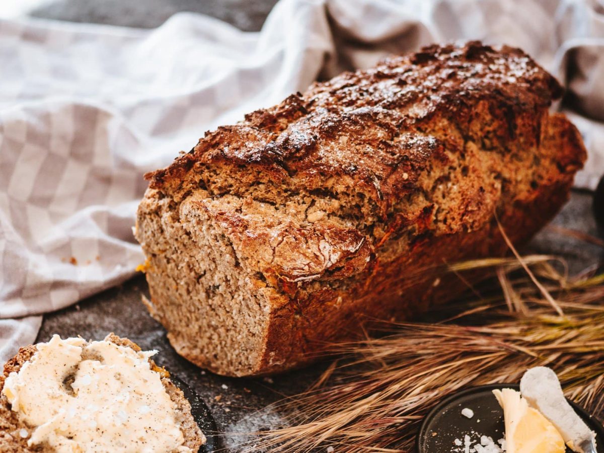 Ein angeschnittenes Dinkelbrot mit Nüssen, daneben liegen Getreideähren, ein Geschirrtuch und Butter.