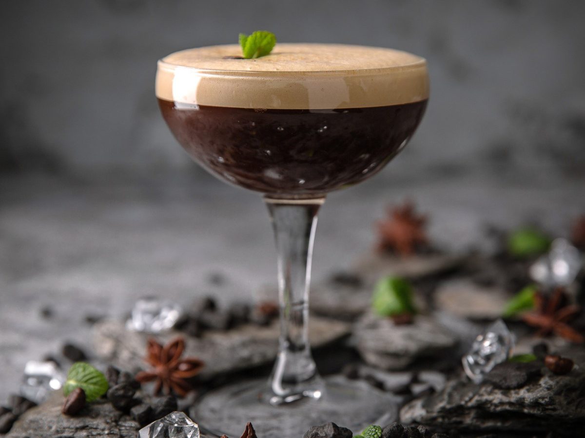Ein Glas Espresso Martini vor dunkelgrauem Hintergrund, drumherum liegen Eiswürfel und Anissterne.