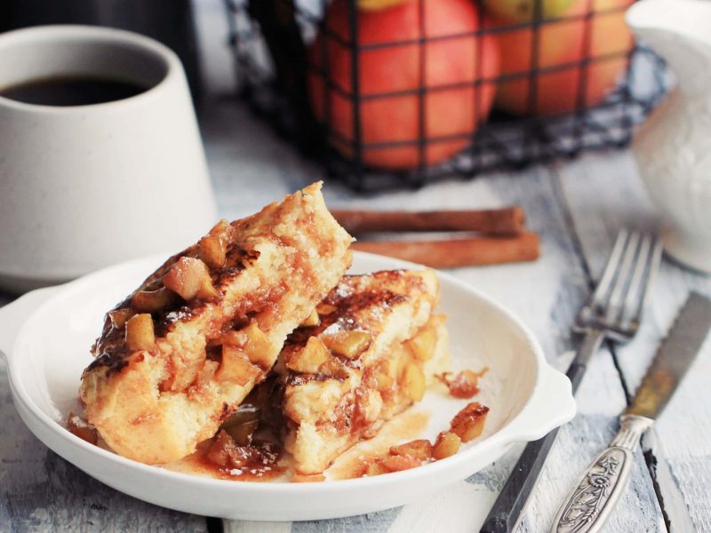 Ein Teller gefüllte French Toasts mit Apfelkompott, daneben Besteck, eine Tasse Kaffee und ein Pott Milch.