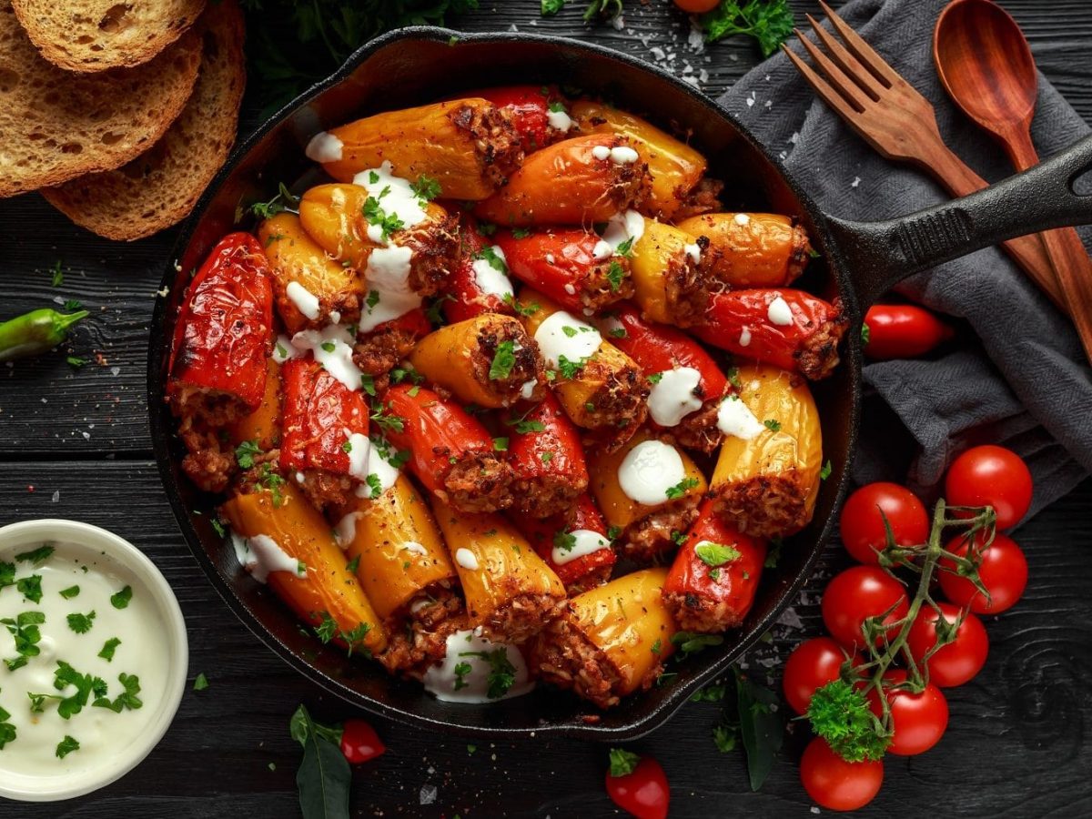 Aus der Vogelperspektive: In einer PFanne sind rote und gelbe Mini-Paprika, die mit Lamm und Feta gefüllt sind. Drumherum liegen Tomaten, ein paar Scheiben Brot, eine SChale mit Joghurt sowie ein Besteck.