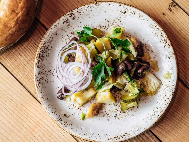 Ein würziger, saftiger georgischer Kartoffelsalat aus Kartoffeln, saurer Gurke, Kidneybohnen, Kohl, Zwiebeln und Petersilie auf einem hellen Tonteller auf hölzernem Untergrund, serviert mit frischem Fladenbrot