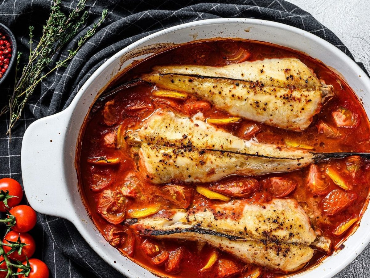 Au der Vogelpersektive: In einem großen Schmortopf liegen drei Seeteufel am Stück, aber ohne Kopf. Seeteufel sind Fische. Diese liegen in einem Bett von Tomaten und Paprika und Rosmarin.