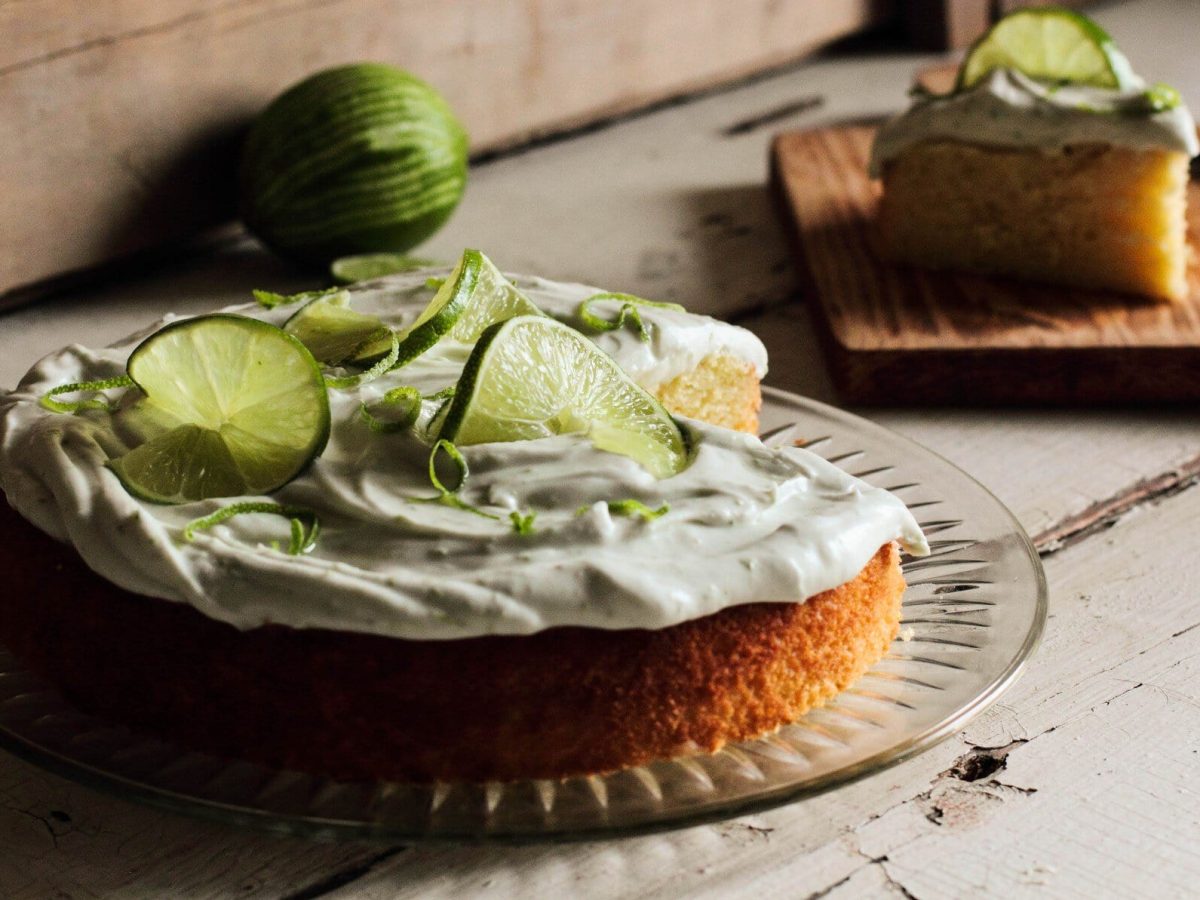 Ein angeschnittener Gin-Tonic-Kuchen auf einer runden Kuchenplatte, dahinter steht ein Stück vom Kuchen auf einem Holzbrett.