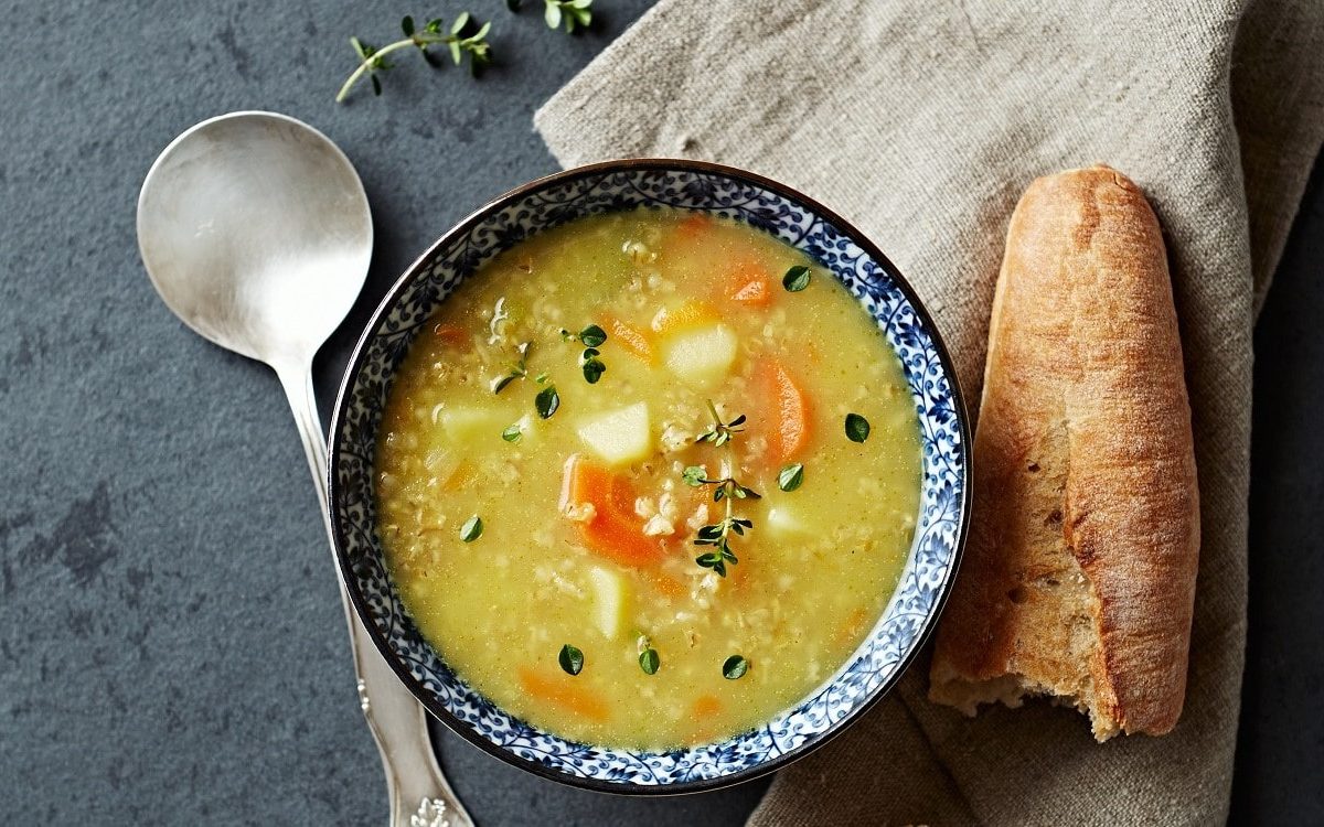 Graupen-Suppe mit Petersilienwurzel, Sellerie, Karotte und Graupen, mit Majoran garniert in einer gemusterten Schale auf einem steinernen Untergrund