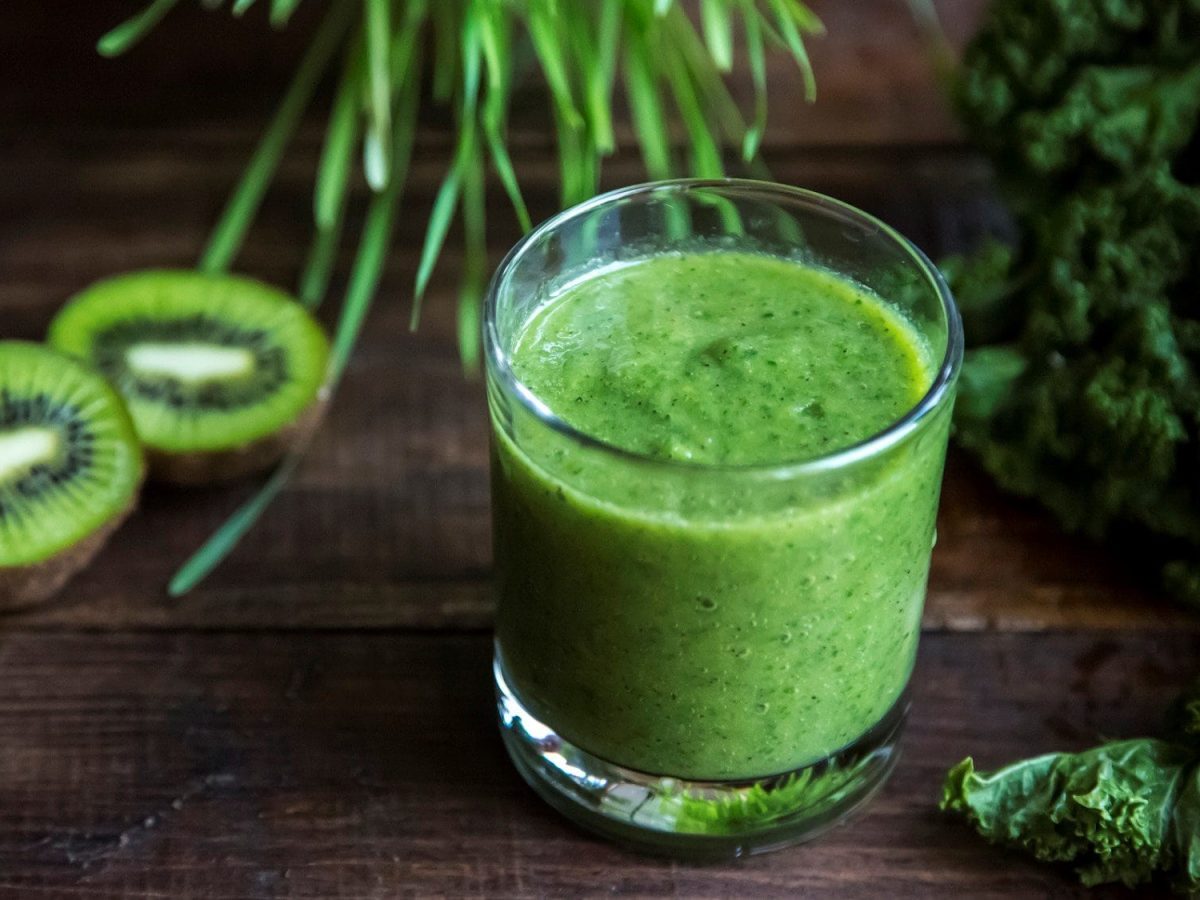 Ein Glas mit Grünkohl-Smoothie. Auf dem dunklen Untergrund liegen Kiwis und Grünkohl.