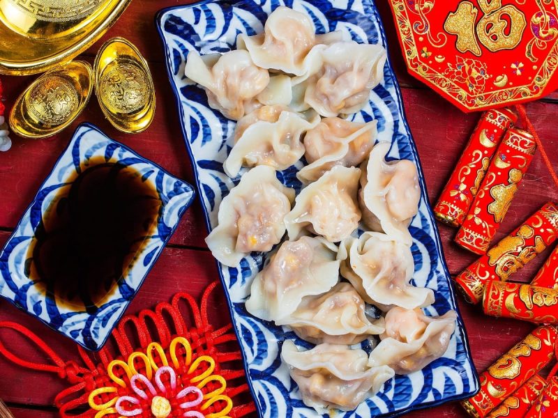 Viele Jiaozi, traditionell chinesische Dumplings auf einem langen Teller angerichtet, neben einer passenden Schale mit Sauce, und vieler roten Neujahrsdekoration vor einem roten Hintergrund, von oben.