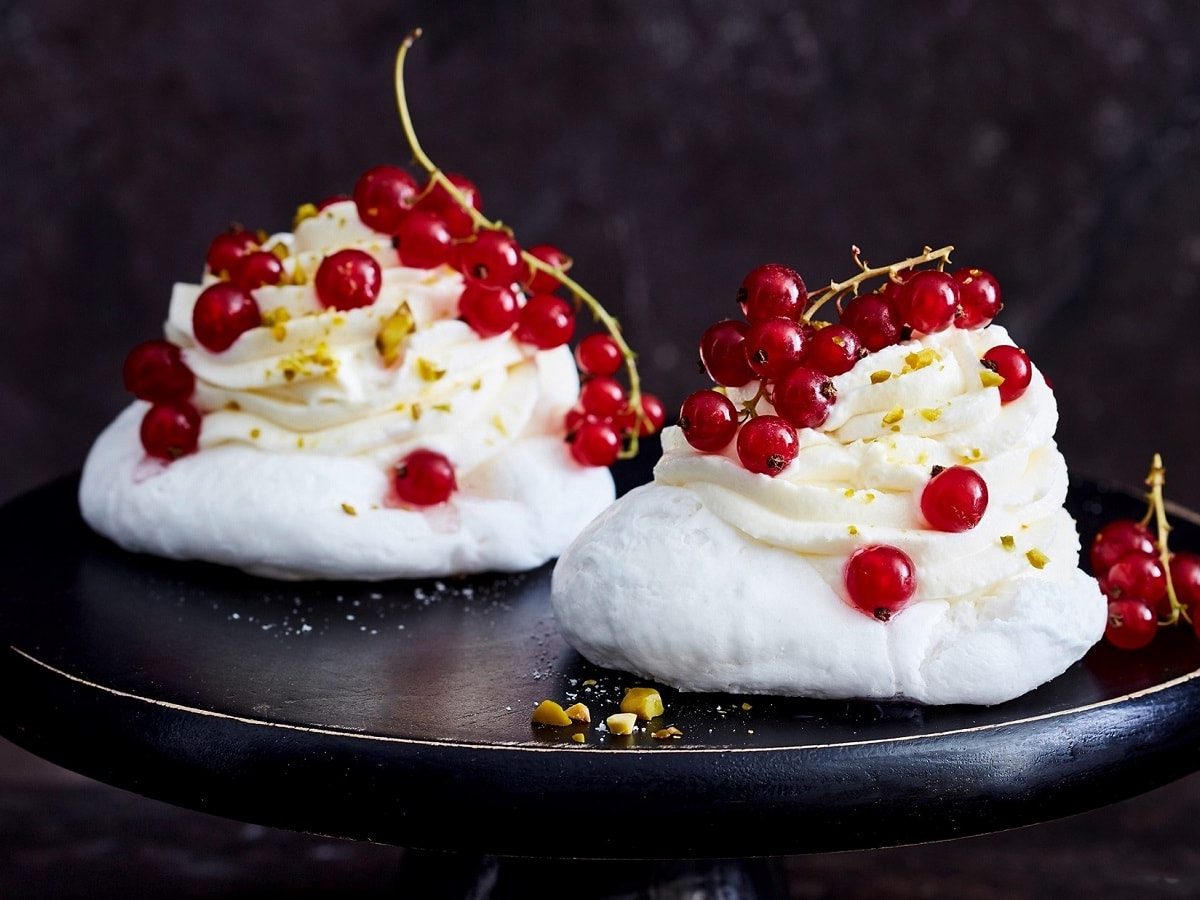 Eine schwarze Platte auf der zwei Johannisbeer-Baisertörtchen garniert mit Pistazien und frischen Johannisbeeren, zu sehen sind, vor einem dunklen Hintergrund.