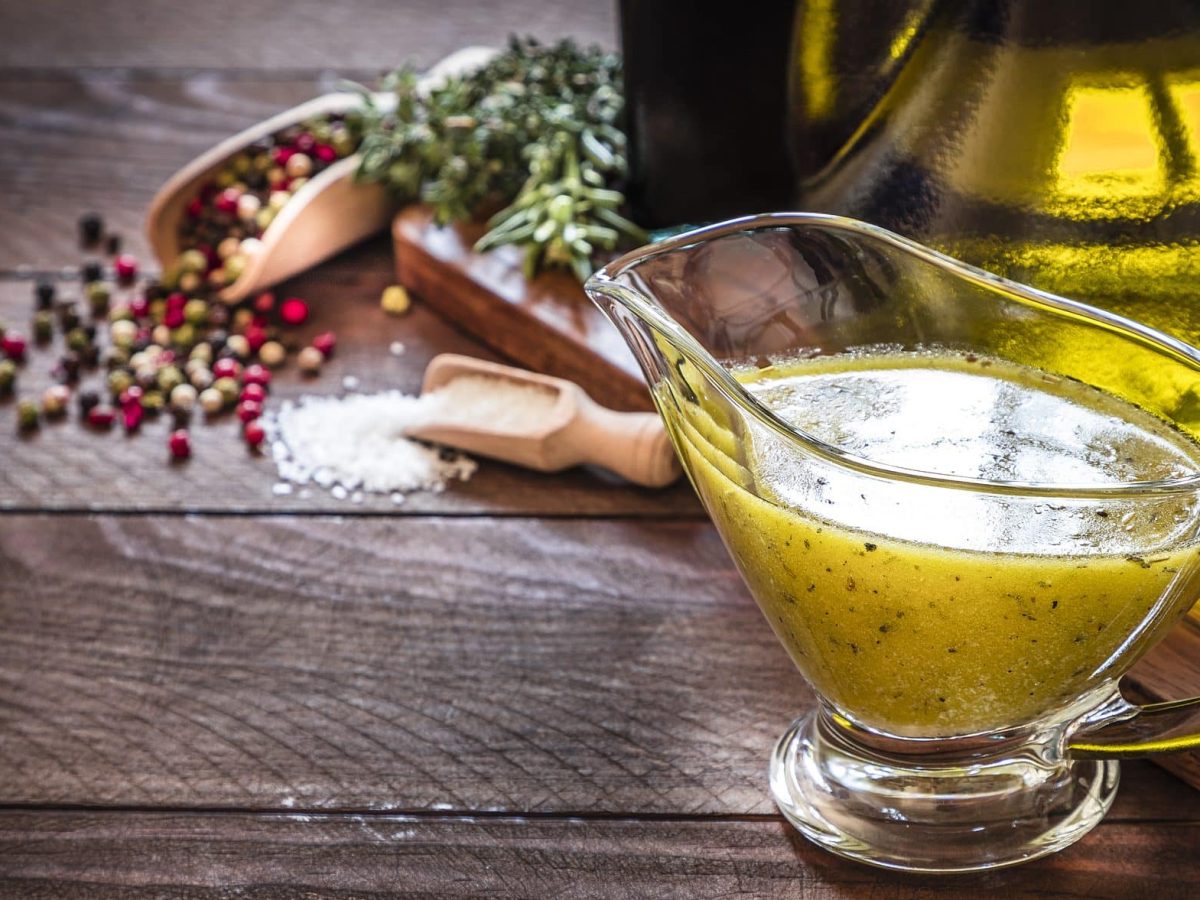 Auf einem Holztisch steht eine Sauciere aus Glas. In dieser ist eine klassische Vinaigrette. Dabei handelt e sich um ein Salatdressing. Zu sehen sind auch eine Glasflasche mit Olivenöl, Pfefferkörner und Kräuter. Sowie ein Holzlöffel mit Salz.