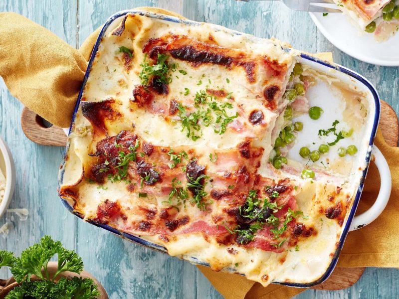 Lasagne alla Carbonara in einer Form mit der cremigen Carbonara-Soße aus Speck, Schmand, Parmesan und Erbsen, serviert auf einem Tuch auf hellem Hintergrund