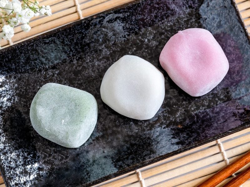 Drei Mochi in verschiedenen Farben, grün, weiß und rosa, auf einem schwarzen, langen Teller, der auf einer Bambusmatte steht, neben einem Strauch weißer Blumen und einem paar Essstäbchen.