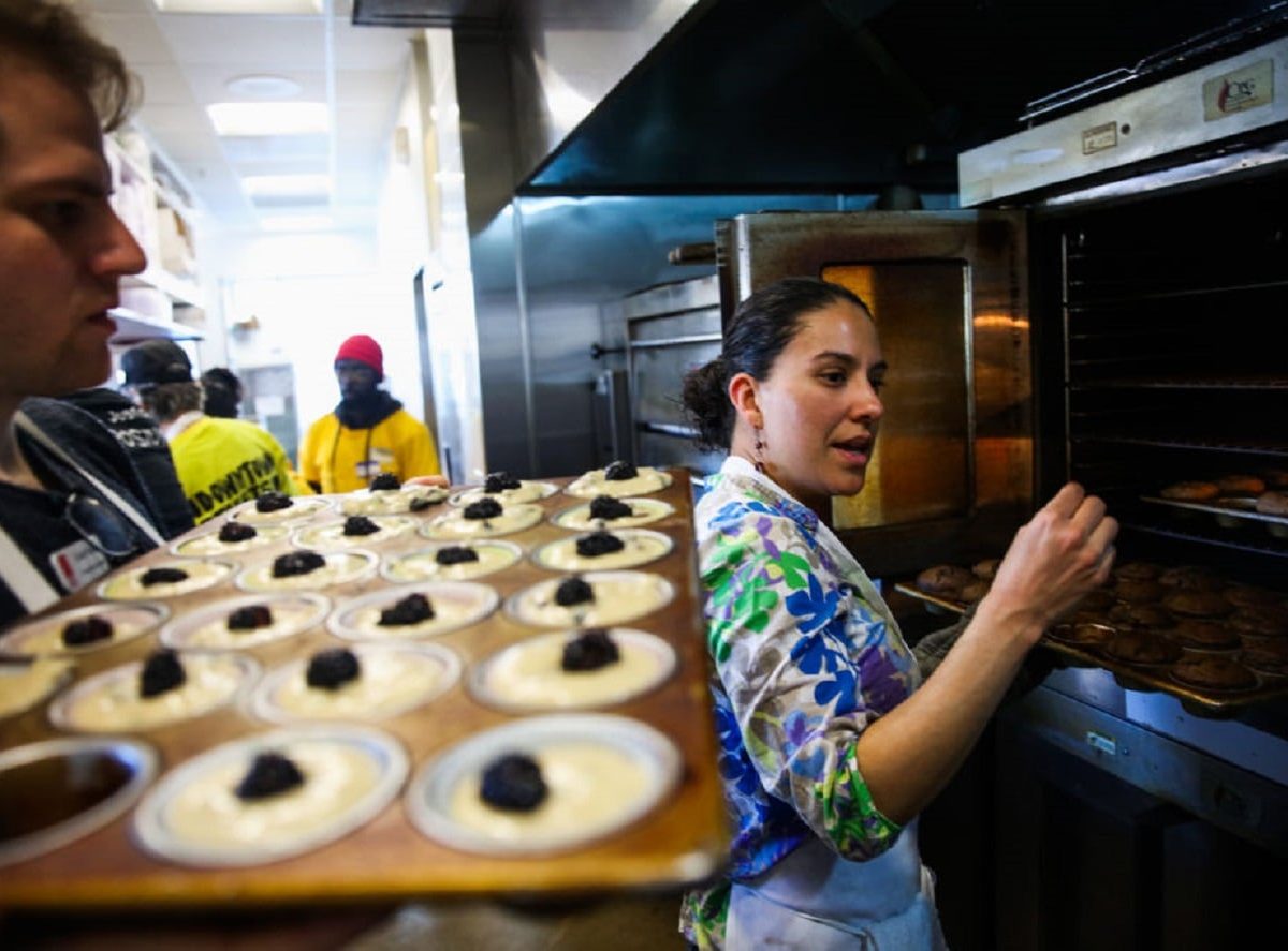 Frau in Profi-Küchen vor geöffnetem Backofen gestikulierend. Mann dahinter hält Backblech Backofen-Zubehör mit Teig. Im Hintergrund Küchenpersonal. Frontalaufnahme.