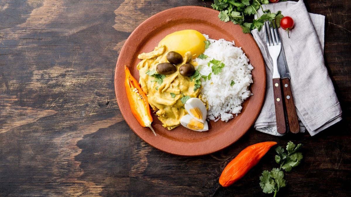 Peruanisches Aji de Gallina, eine Art Hühnercurry nach peruanischem Rezept mit Reis, Kartoffel, Oliven und frischer Chili in Tonschale serviert auf steinernem Untergrund