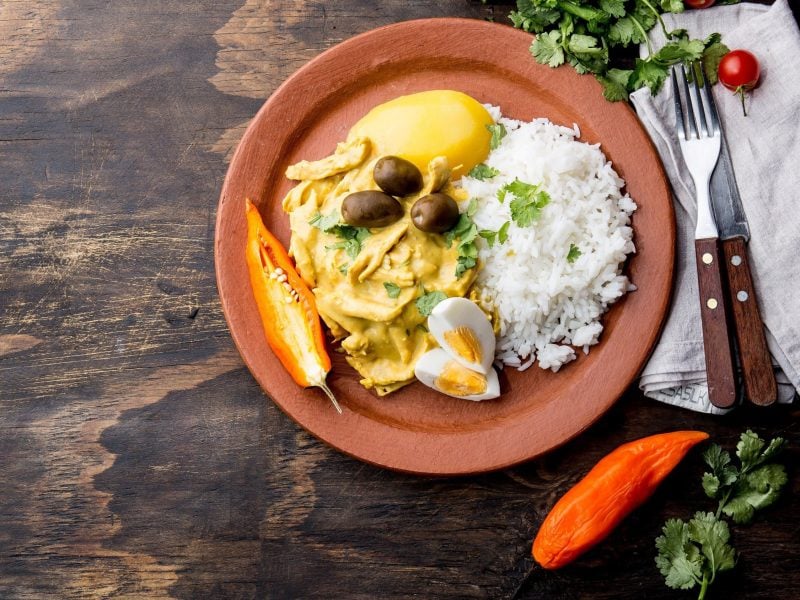 Peruanisches Aji de Gallina, eine Art Hühnercurry nach peruanischem Rezept mit Reis, Kartoffel, Oliven und frischer Chili in Tonschale serviert auf steinernem Untergrund