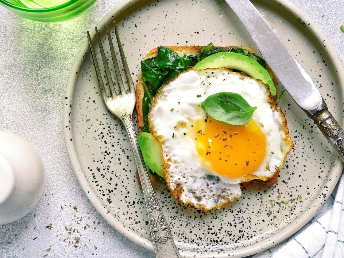 Pesto Eggs auf einem Frühstückstoadt mit Spinat und Avocado. Neben dem Toast liegen Messer und Gabel auf einem gemusterten Teller.