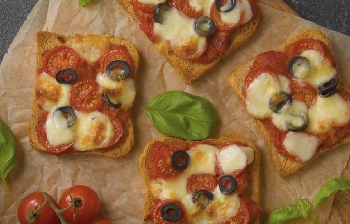 Vier Stücke von unserem Pizza-Toast liegen mit Basilikum und Tomaten garniert auf einem Holzbrett.