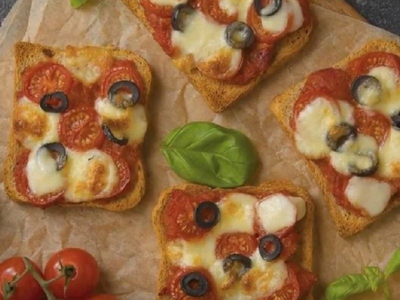 Vier Stücke von unserem Pizza-Toast liegen mit Basilikum und Tomaten garniert auf einem Holzbrett.