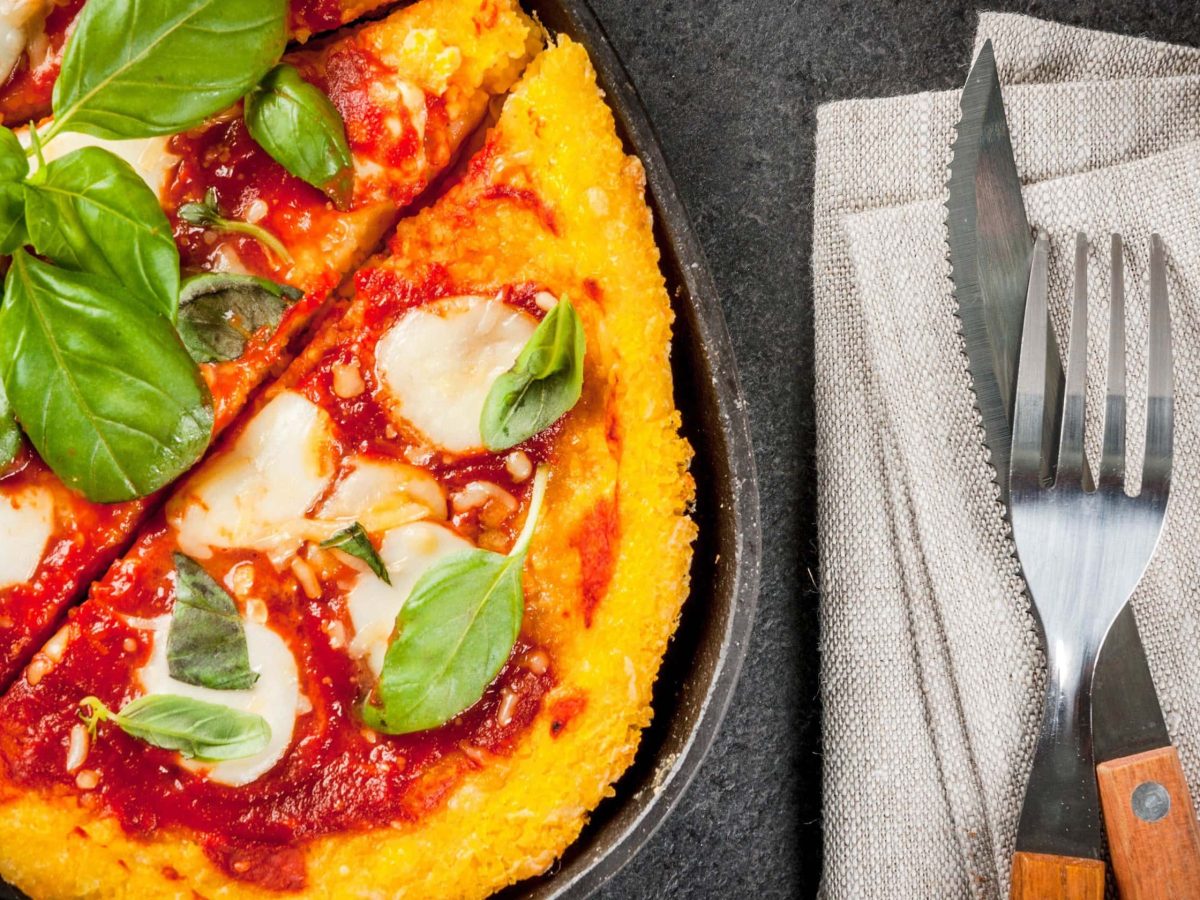 Saftige Polenta-Pizza belegt mit Tomatensoße und Mozzarella, garniert mit frischem Basilikum; serviert in einer heißen Pfanne auf grauem Holzhintergrund