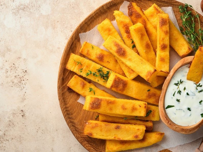 Kross gebackene Polenta-Schnitten mit Kräutern bestreut und dazu frischer Dip serviert auf einem hölzernen Teller auf hellem Hintergrund