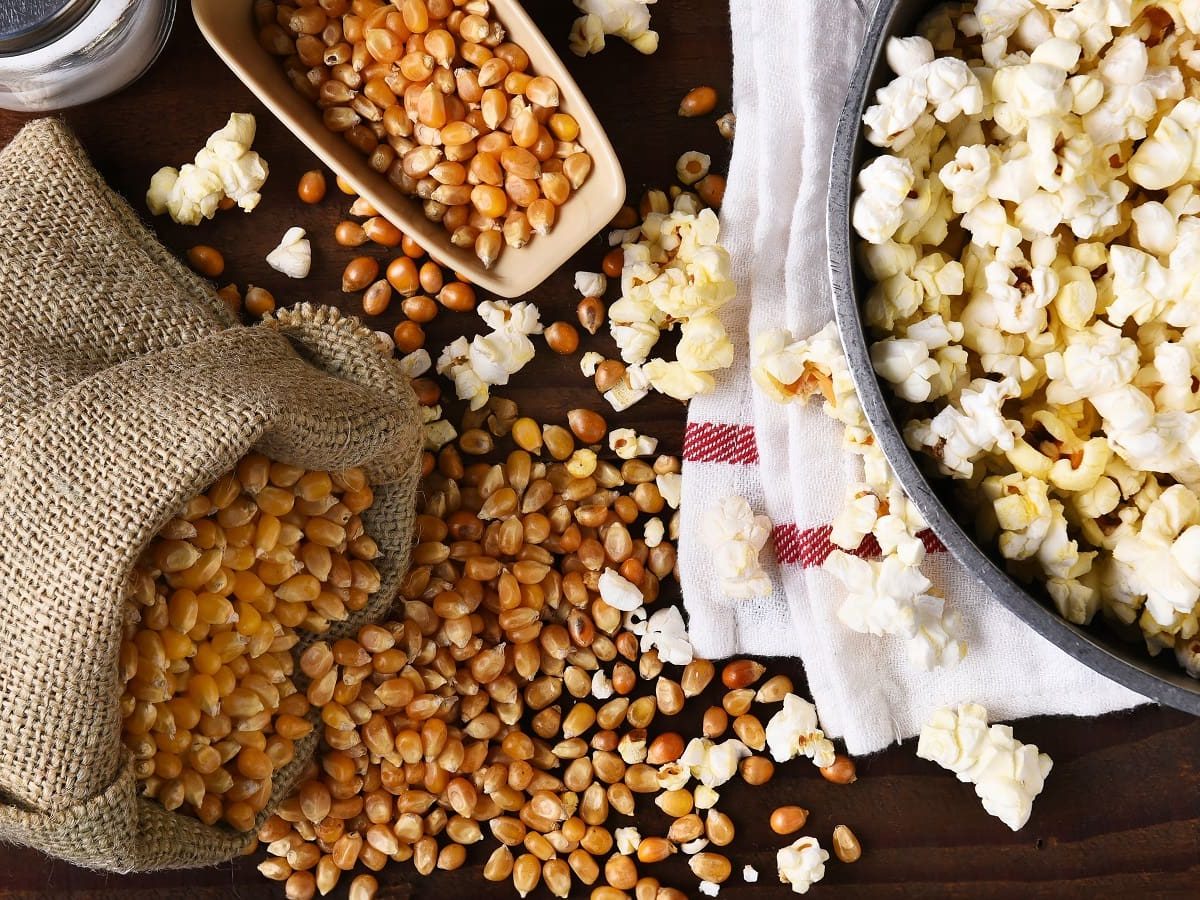 Der Prozess von Popcorn selbst machen: Links im Bild ist ein kleiner Sack mit noch ungepoppten Körnern, die sich auf dem Tisch mit bereits gepoppten vermischen, während rechts im Bild ein Topf mit gepoppten Popcorn zu sehen ist, alles vor einem Holzhintergrund, von oben.