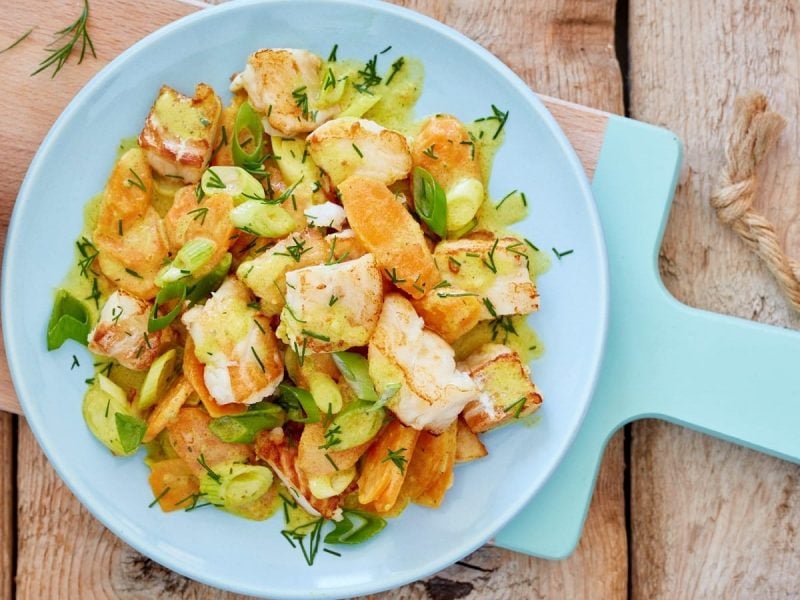 Ein hellblauer Teller auf einem Holz-Schneidebrett, auf dem eine Portion Ragout mit Seelachs und Karotten angerichtet ist, das mit Dill überstreut ist. Neben dem Teller steht eine kleine Schüssel mit Dill. Alles ist vor einem Holzhintergrund von oben fotografiert.