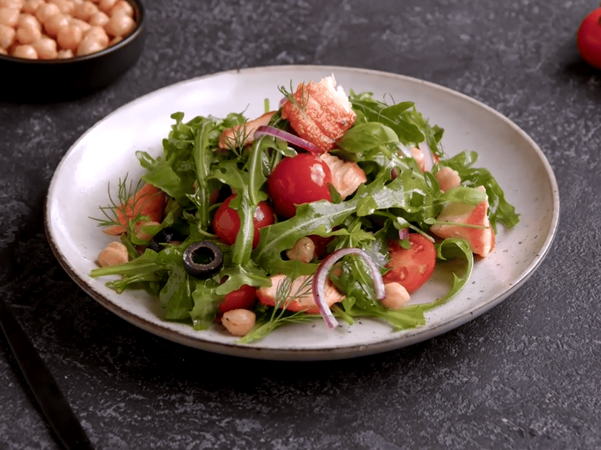 Ein Teller Rucolasalat mit Lachs und Kichererbsen, außerdem Tomaten, roten Zwiebeln und Oliven. Der Teller steht auf dunklem Untergrund, daneben eine Gabel, Tomaten und eine Schale Kichererbsen.